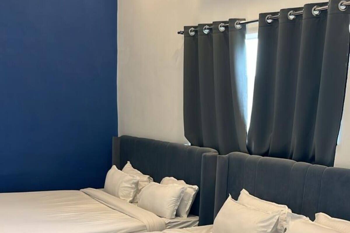 Een tweepersoonsbed met witte lakens en een donker hoofdbord, voor een blauwe muur met een raam en gordijnen.