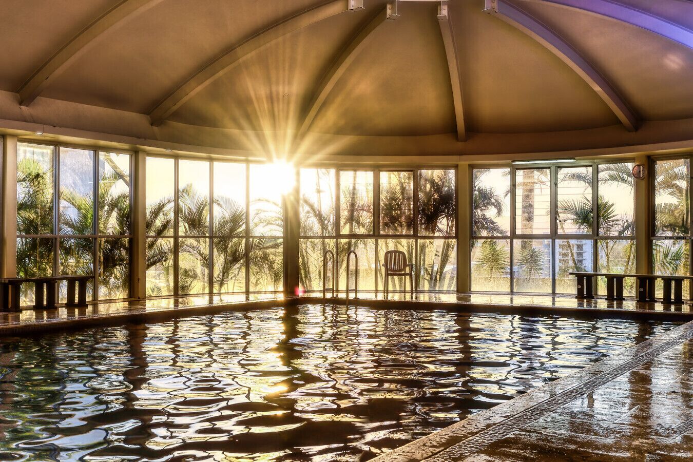 Une vaste piscine intérieure, baignée de lumière naturelle, entourée de grandes fenêtres qui offrent une vue sur les palmiers.