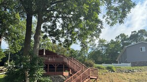 A wooden dock leading to a house with a deck and stairs, surrounded by trees and a clear pond.