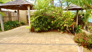 A paved walkway leading to a covered area with thatched roofs, surrounded by lush greenery and plants. - Lakeside Getaway - 4BR Jetty Wi-fi Waterfront (Encounter Bay)