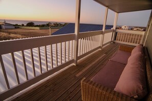 A covered porch with a view of the ocean, wooden flooring, and a wicker chair with a cushion.