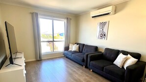 Ein Wohnzimmer mit Fernseher auf einem weißen Schrank, einem dunkelgrauen Sofa mit Kissen, einem Fenster mit Vorhängen und einer Klimaanlage an der Wand.
