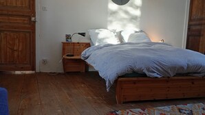 A bedroom with a wooden floor, a bed with a patterned blanket, a bedside table with a lamp, and a mirror on the wall.