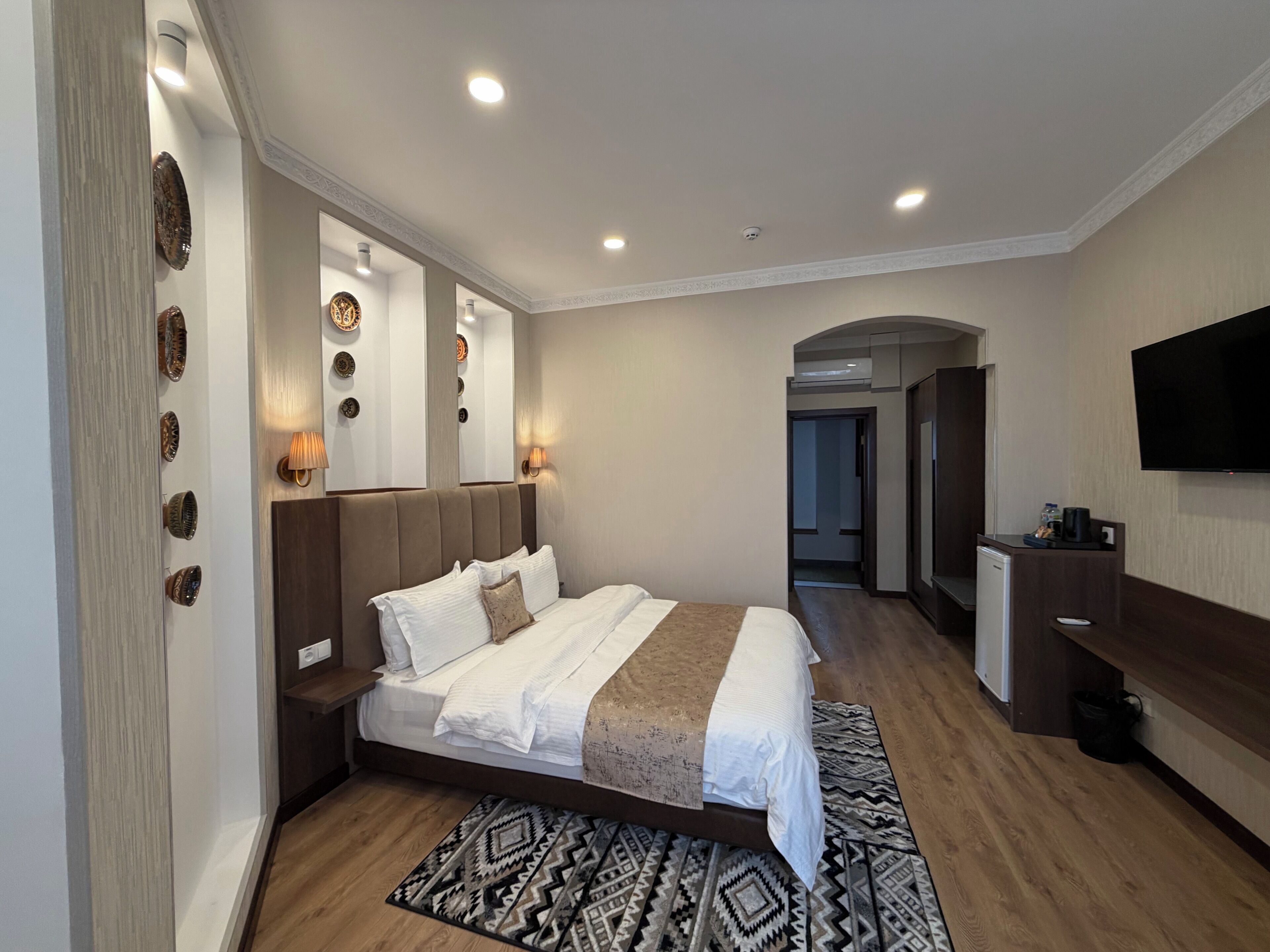 A modern hotel room with a large bed, bedside tables, a flat-screen TV, and a mirror.