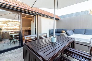 Un patio al aire libre con una mesa y sillas de madera, un cojín azul y vistas de la ciudad a través de un ventanal.