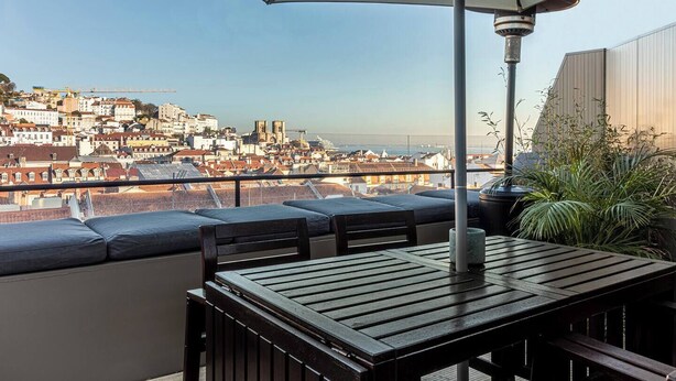 Un balcón con mesa y sillas, un paraguas grande y vistas a un paisaje urbano con edificios y una masa de agua.