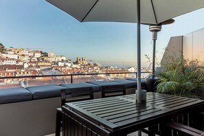Un balcón con mesa y sillas, un paraguas grande y vistas a un paisaje urbano con edificios y una masa de agua.
