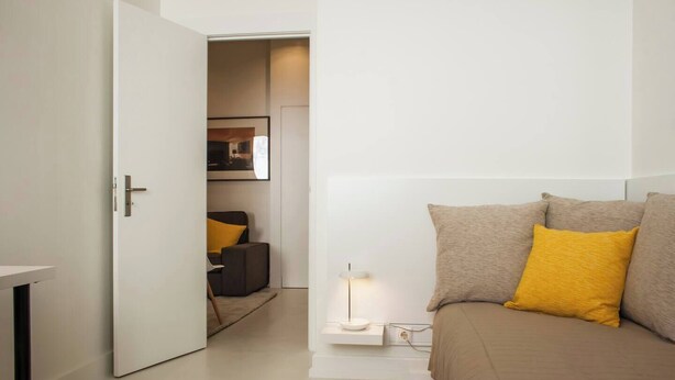 A modern hotel room with a bed, a bedside table, a lamp, and a door leading to a hallway.