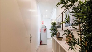 A corridor with a white door, a white refrigerator, and a fire extinguisher. There are plants and framed pictures on the wall.