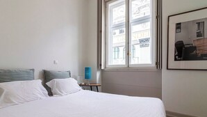 A hotel room with a large bed, a small table with a lamp, and a window with a view of buildings.