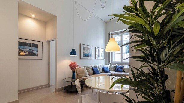 A modern living room with a round dining table, white chairs, a sofa with blue and beige cushions, and a large green plant.