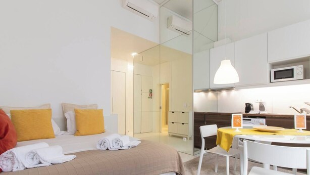 A modern hotel room with a bed, a dining area, and a kitchenette.