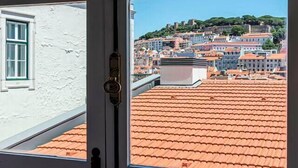 Una veduta attraverso una finestra mostra un edificio con tetto in tegole rosse, pareti bianche e un balcone con ringhiera in metallo.