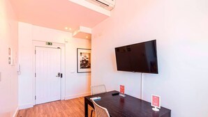 Una camera d'albergo con pavimento in legno, un tavolo da pranzo nero, una TV fissata al muro e una porta con un cartello sopra.