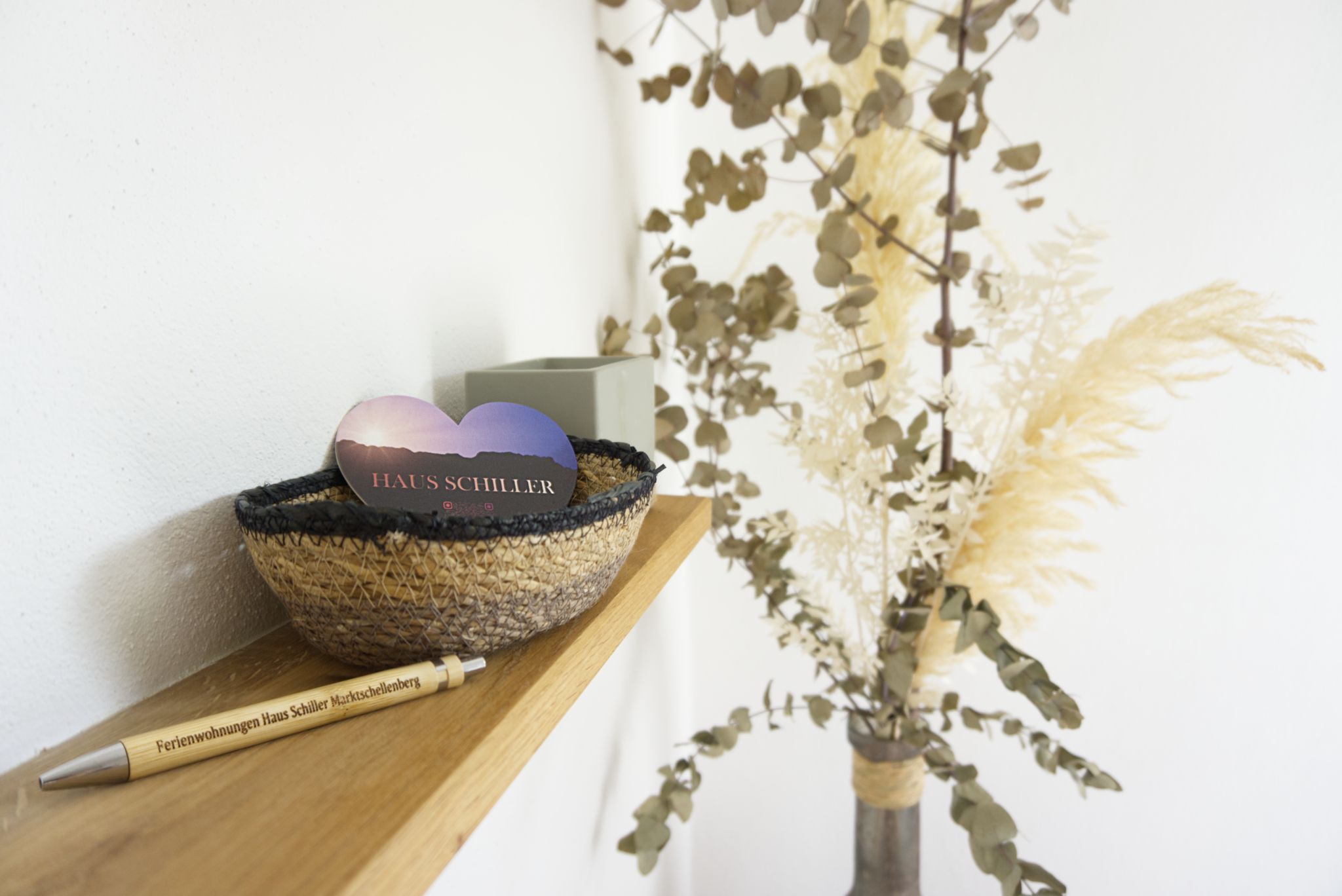 A woven basket with a heart-shaped object on a wooden shelf, next to a pen and a vase with dried plants.