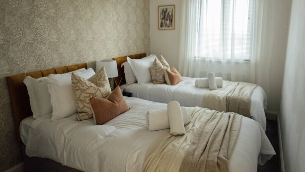A hotel room with two beds, each with white linens and multiple pillows. There is a large window with sheer curtains, a framed picture on the wall, and a woven headboard. - Greenfields House Ashford (Kent)