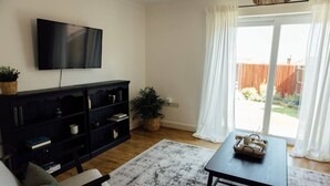 A living room with a flat-screen TV, a dark wooden bookshelf, a light-colored sofa, a patterned rug, and a table with a basket. - Greenfields House Ashford (Kent)