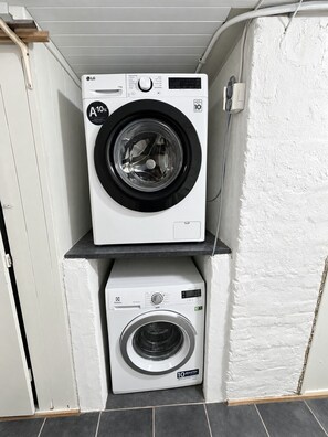 Deux machines à laver superposées dans un coin, avec un mur blanc et une porte en bois.