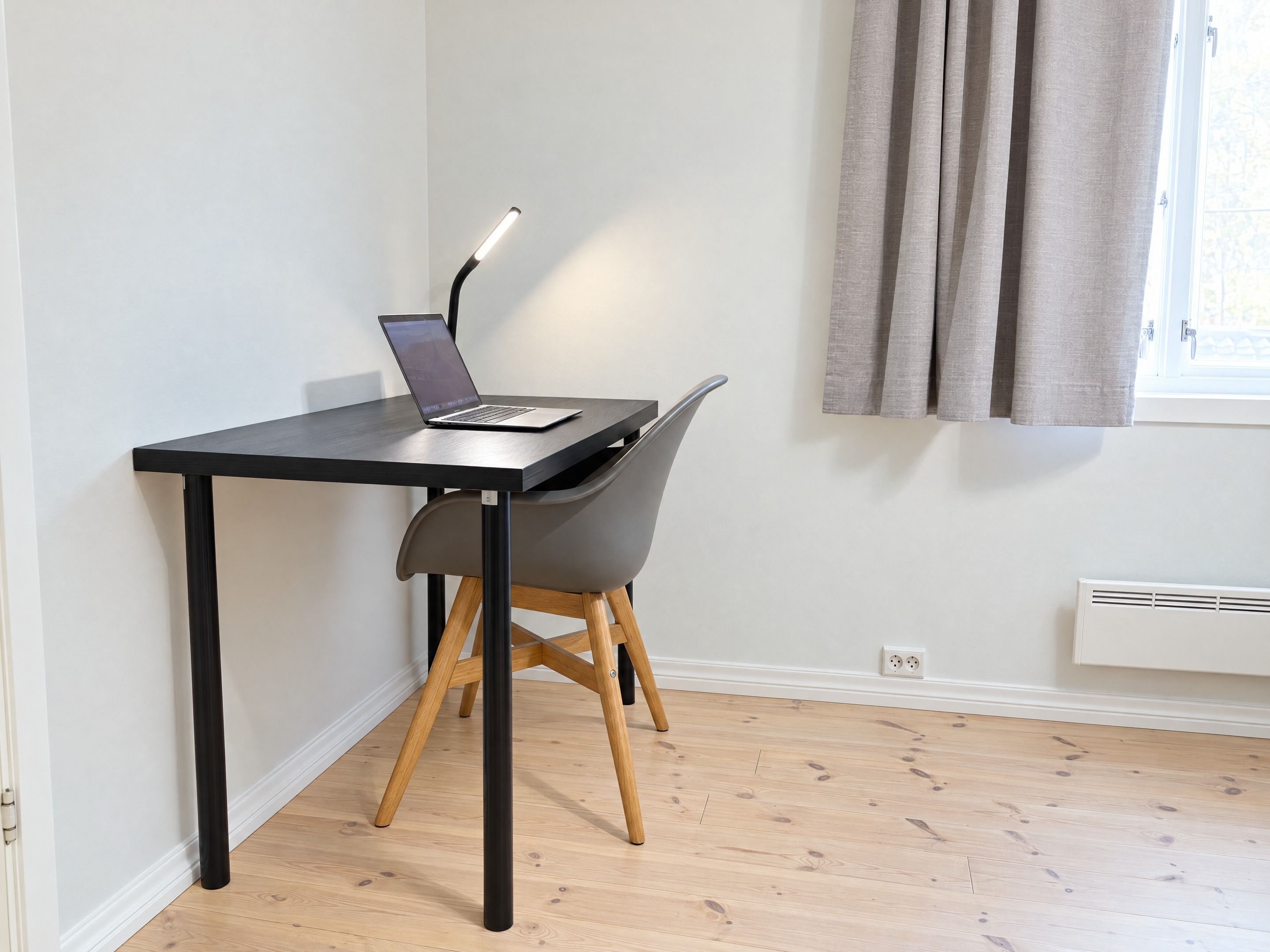 A modern workspace with a laptop on a black table, a grey chair, and a window with curtains.