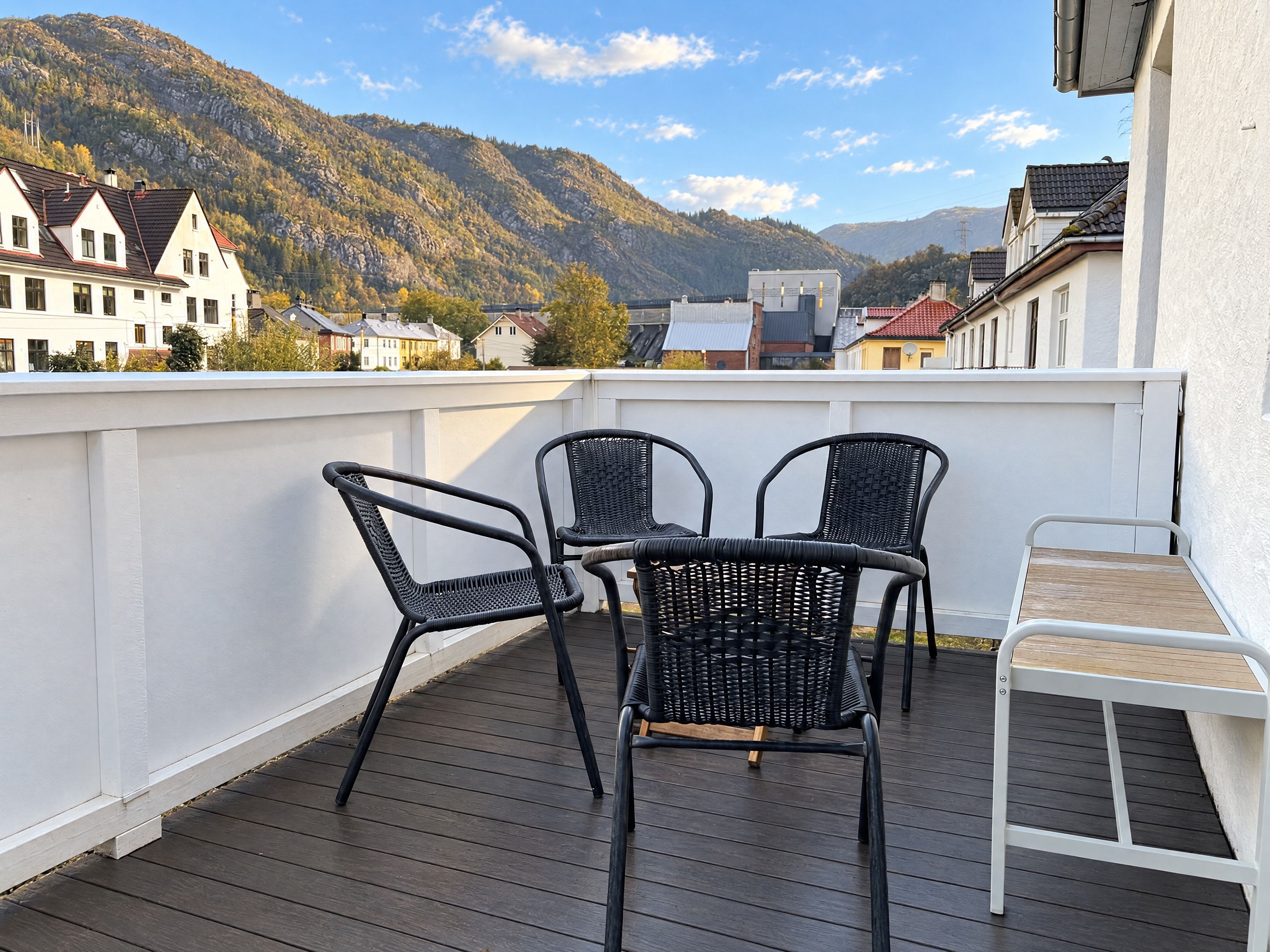 A balcony with two wicker chairs and a small table, overlooking a mountainous landscape with houses in the background.