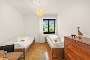A room with two single beds, a wooden dresser, a desk with a chair, and a window with a view of greenery. - Casa Ana - Losone Center (Losone)