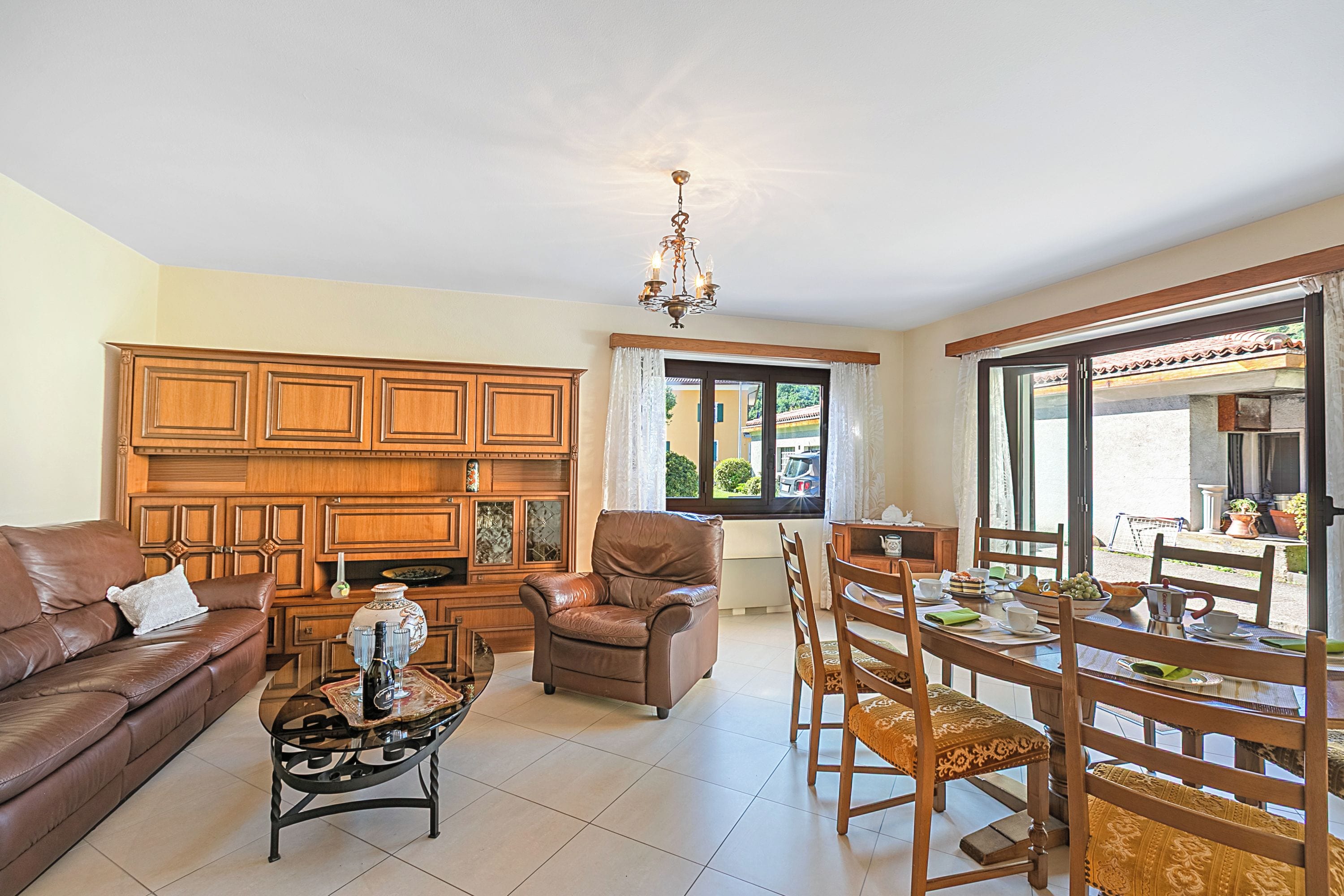 A living room with a brown leather sofa, a brown leather armchair, a wooden dining table with chairs, and a chandelier.