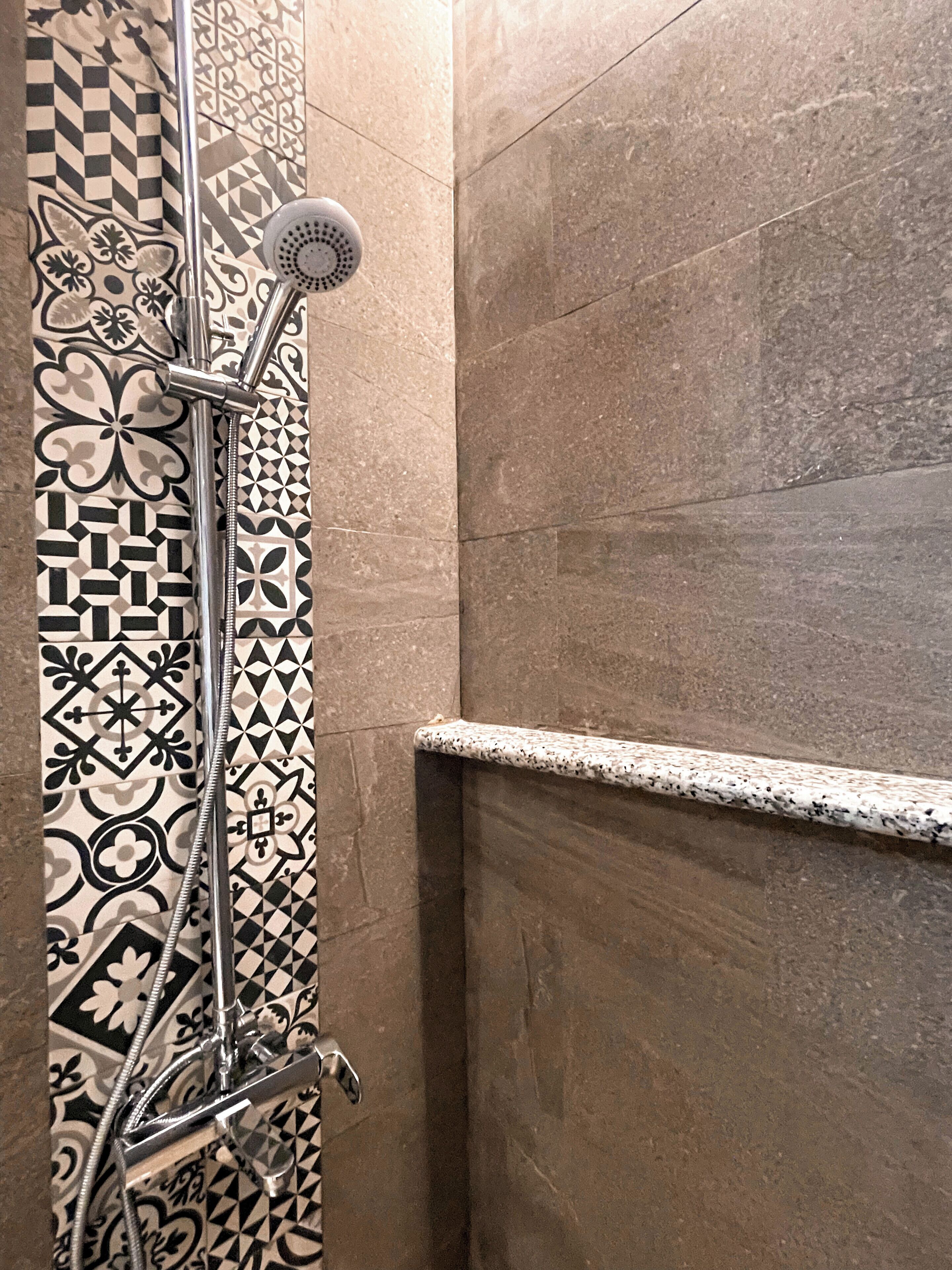 A shower with a handheld showerhead and a wall-mounted control valve. The shower area has a tiled wall with a geometric pattern.