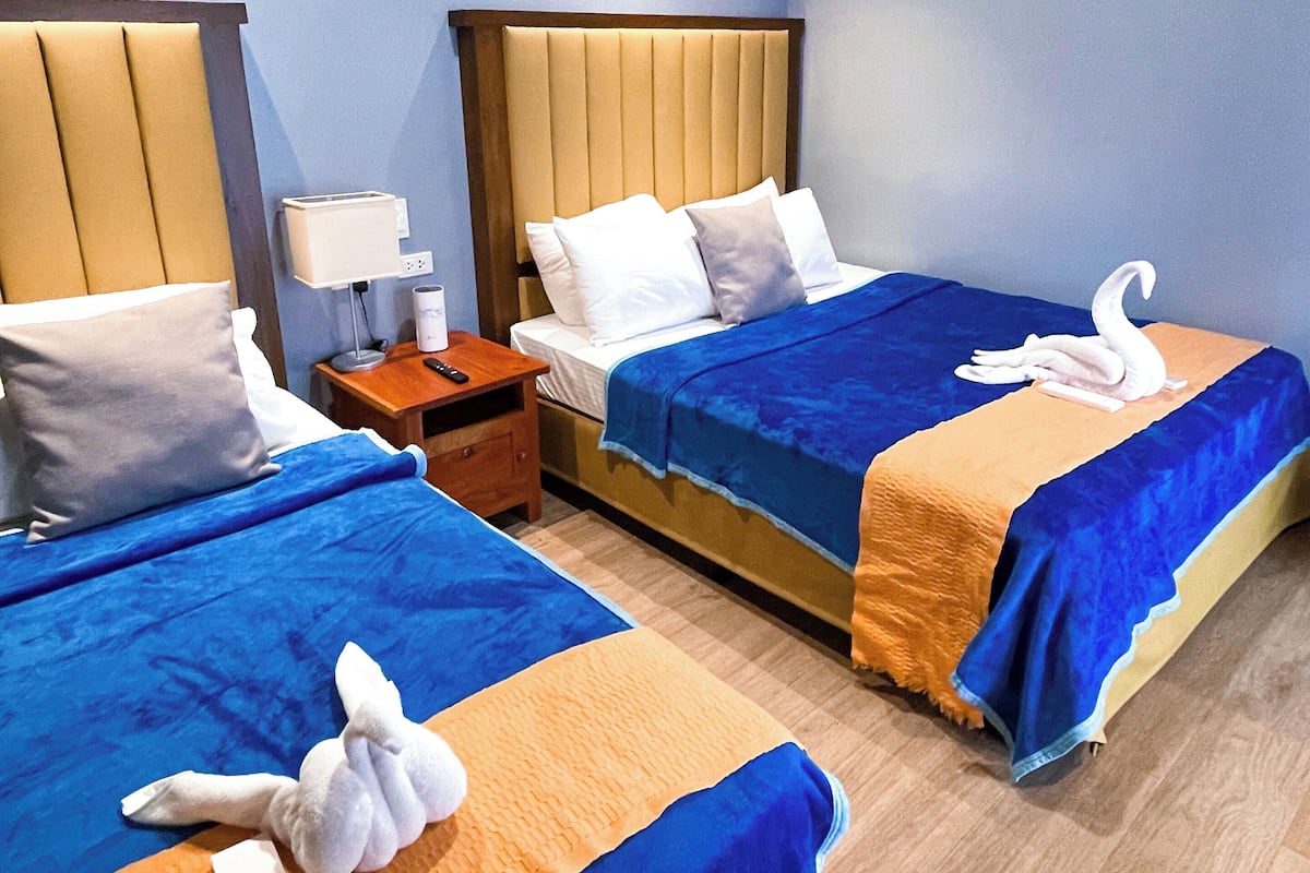 A hotel room with two beds, each with a blue and orange bedspread, and a wooden headboard. There is a nightstand with a lamp and a remote control.