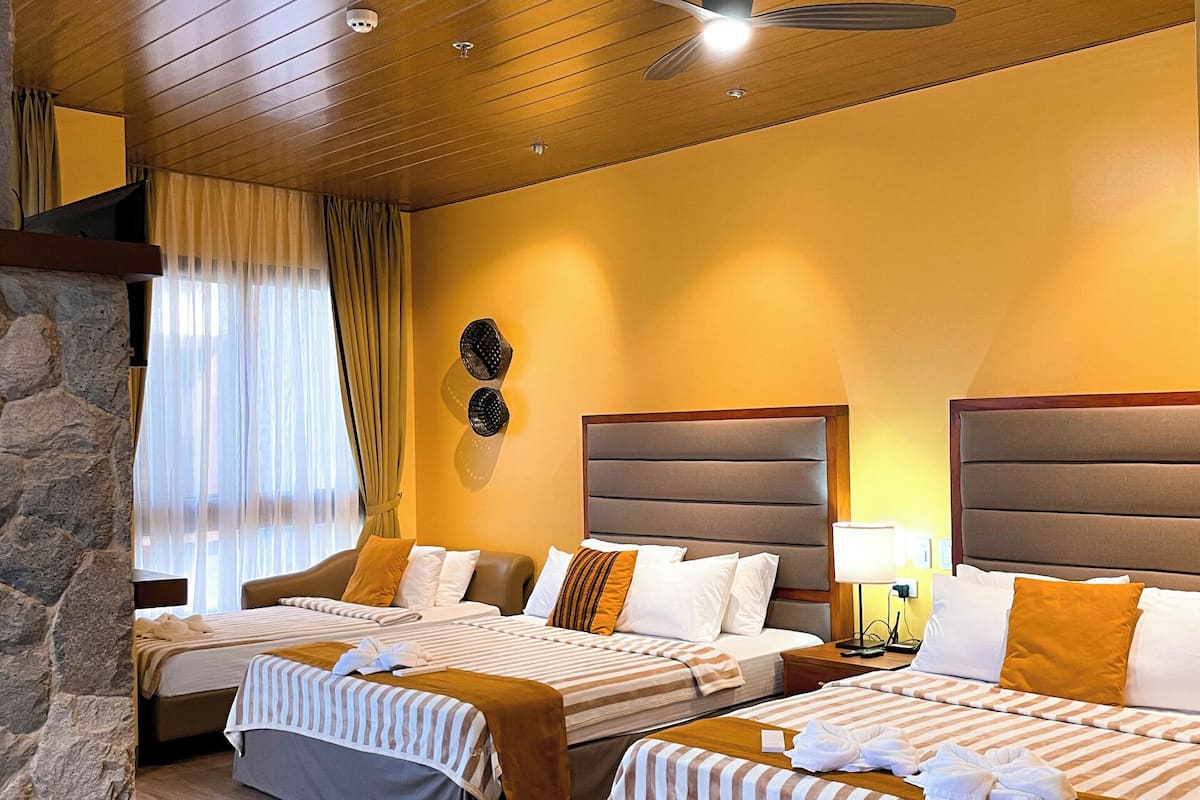 A hotel room with three beds, wooden ceiling, stone wall, and a large window with curtains.