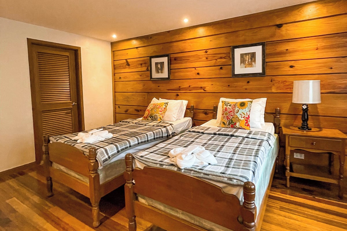 A room with two wooden beds, a wooden wall panel, a wooden door, and a nightstand with a lamp.