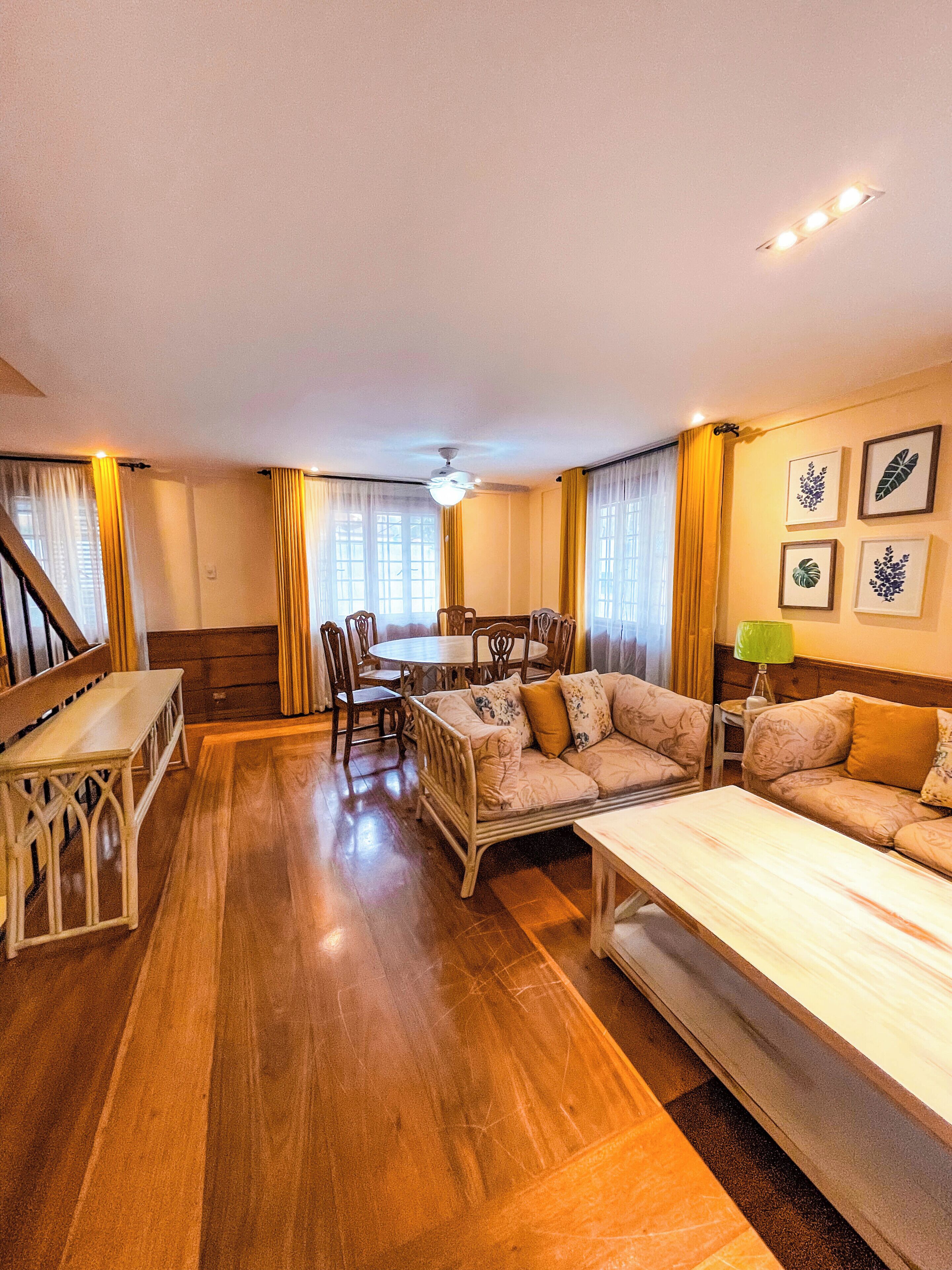 A spacious room with a wooden floor, a sofa set, a dining table with chairs, and a ceiling fan.