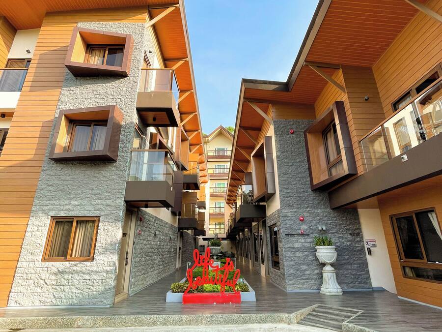 Un pasillo moderno de hotel con varios balcones, un jardín central con un letrero rojo y una pared con textura de piedra.