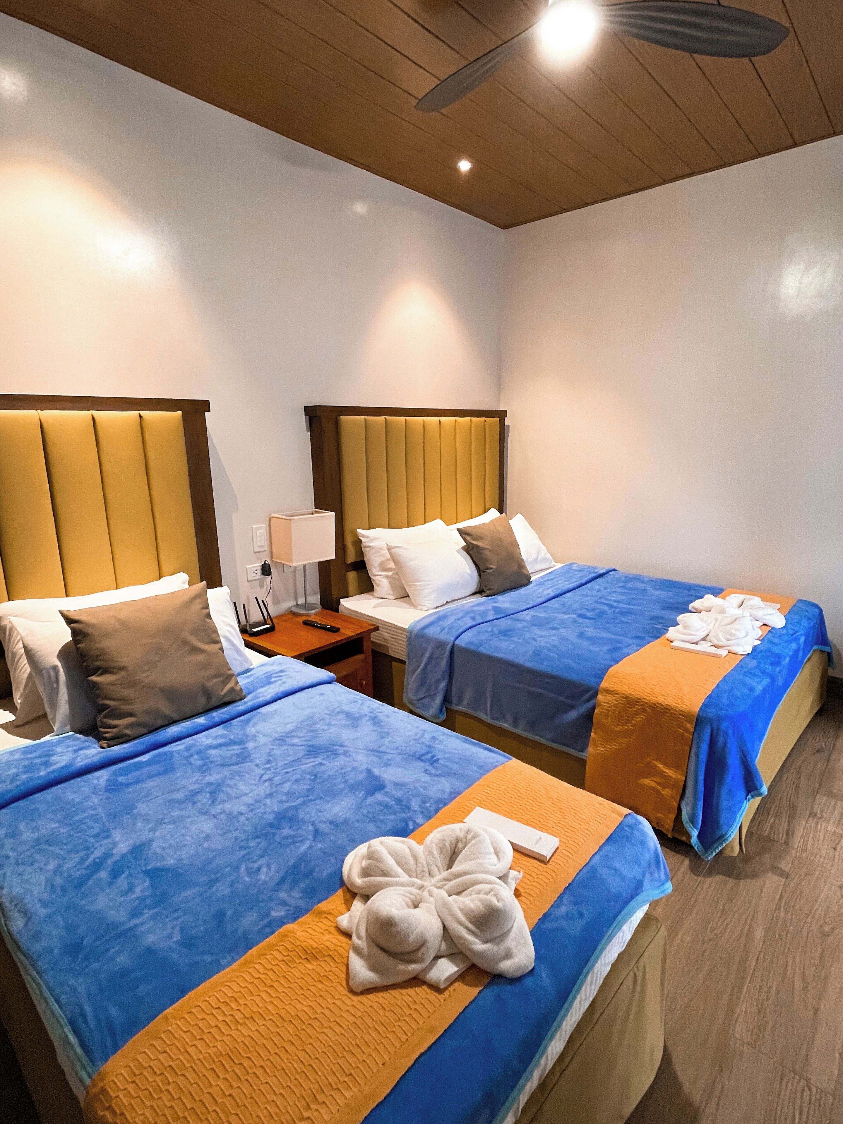 A hotel room with two beds, each with a blue bedspread and an orange blanket. There are white towels on the beds, and a nightstand with a lamp and a remote control.