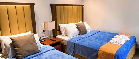 A hotel room with two beds, each with a blue bedspread and an orange blanket. There are white towels on the beds, and a nightstand with a lamp and a remote control.