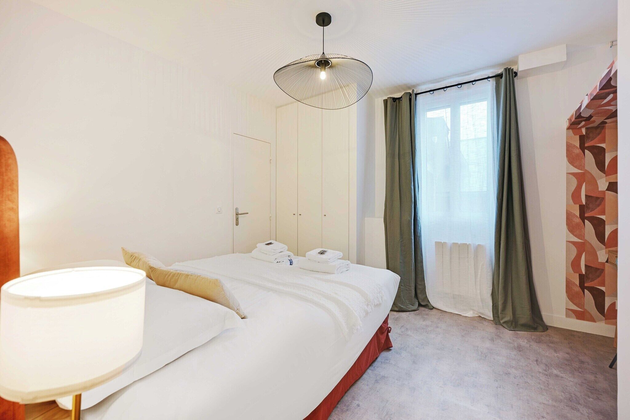Habitació d'hotel amb llit, làmpada de nit, finestra amb cortines i paret amb un dibuix geomètric.