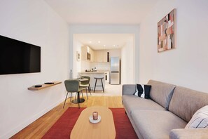 A modern living room with a grey sofa, a wooden coffee table, and a red rug. There is a kitchen in the background with a fridge, microwave, and a dining area with chairs. - Cosy appartement - 1BDR/4P - Boulogne Billancourt (Boulogne-Billancourt)