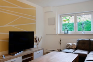 En moderne stue med et fladskærms-tv, et træ sofabord, en brun lædersofa og et vindue med udsigt over grøn bevoksning.