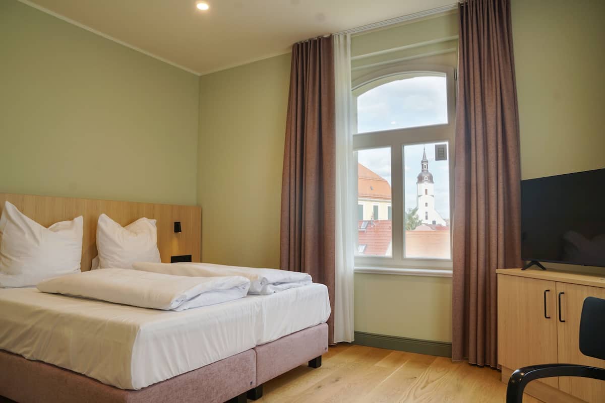 A hotel room with a bed, a television, a window with curtains, and a view of a building outside.