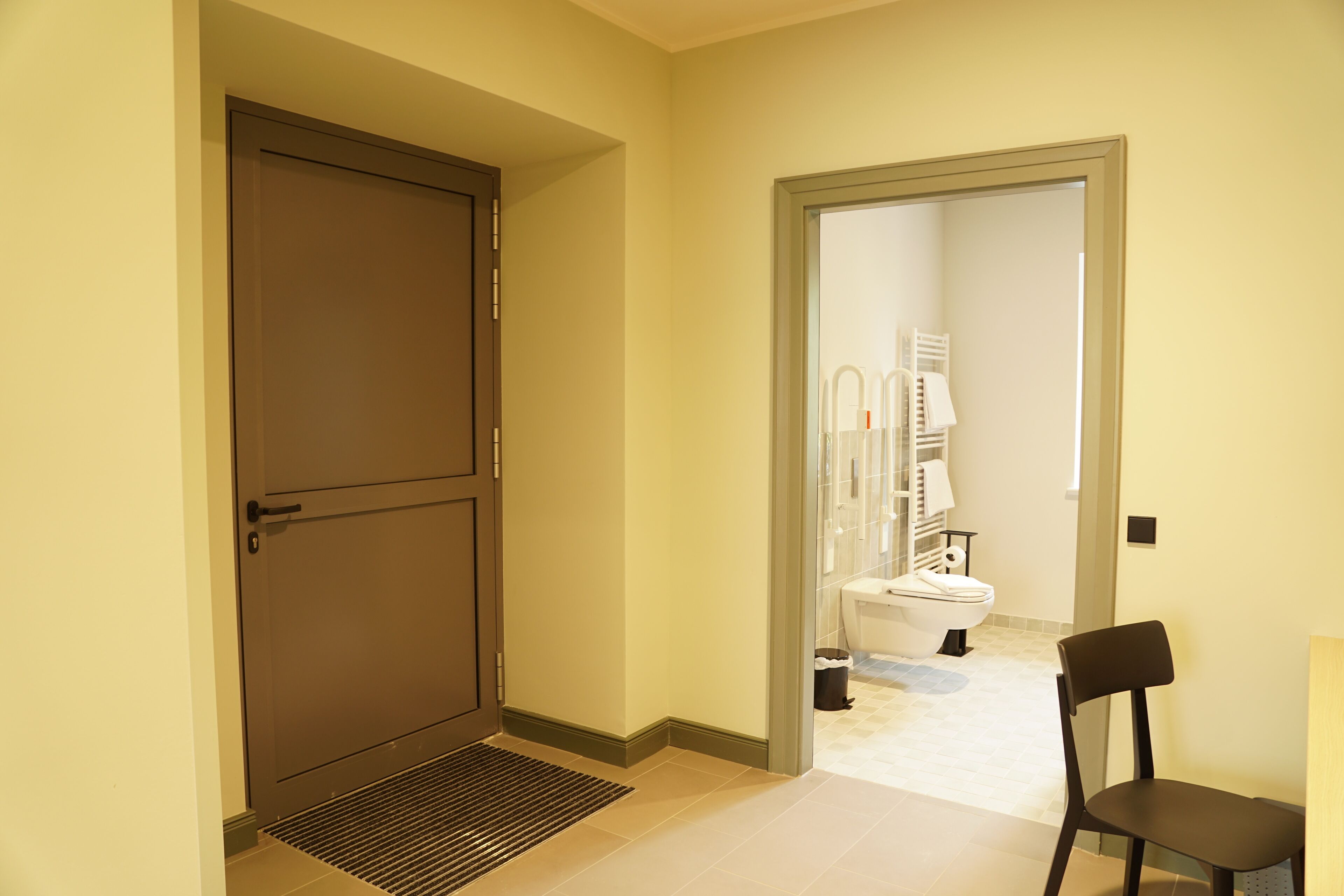 A hallway leading to a bathroom with a toilet, a chair, and a door.