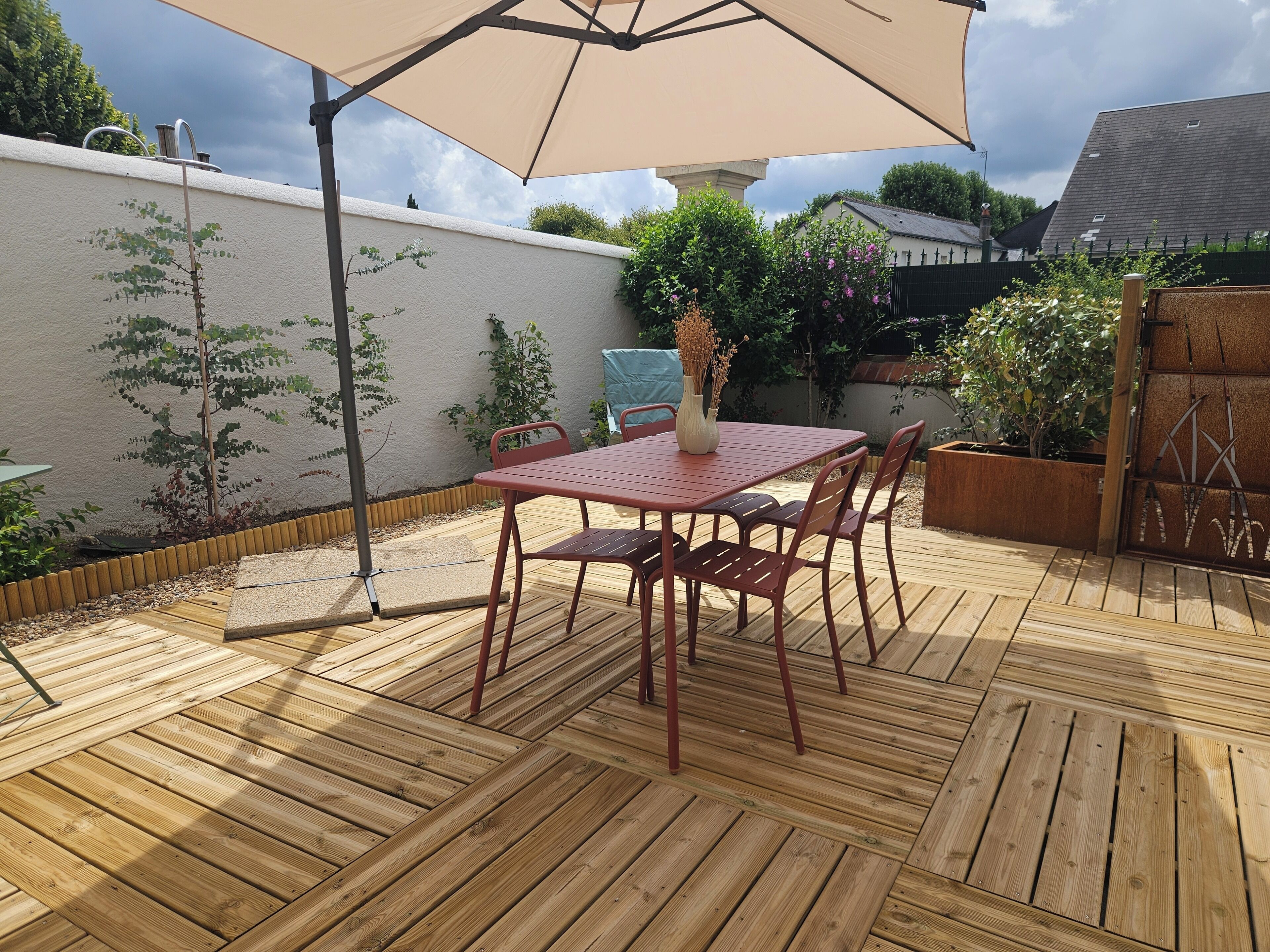 Una terraza de madera con una mesa y sillas rojas, un paraguas y plantas en macetas.