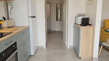 A modern kitchen with a refrigerator, oven, and microwave. There is a wooden countertop with a sink and a small table with a kettle and cups. A door leads to a hallway and another room.