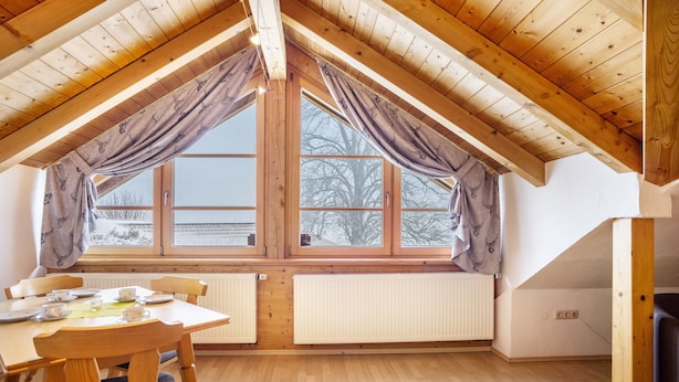 A room with a sloped wooden ceiling, a dining table with chairs, a window with curtains, and a radiator.