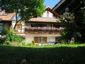 A two-story building with a balcony adorned with flowers, a visible window with a plaque, and a bird feeder.
