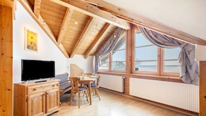 A room with a wooden ceiling, a TV on a wooden cabinet, a dining table with chairs, and large windows with curtains.