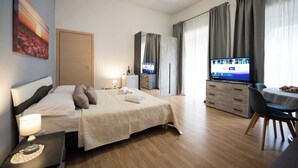 A hotel room with a bed, bedside table, lamp, TV, and a dining area with a table and chairs. - HOME Joséphine, a stone's throw from the Palace, Garden,parking,Kitchen, (Caserta)