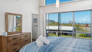 Een slaapkamer met een groot bed, een houten dressoir, een spiegel en uitzicht op de omgeving door grote ramen.