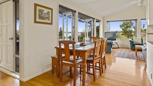 Un comedor espacioso con una mesa y sillas de madera, ventanas grandes que ofrecen vistas al exterior y una zona de estar acogedora con sofá y televisor.