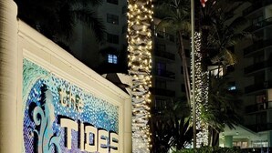Une enseigne portant l’inscription « The Tides » et un motif décoratif, le tout entouré de verdure et de palmiers.