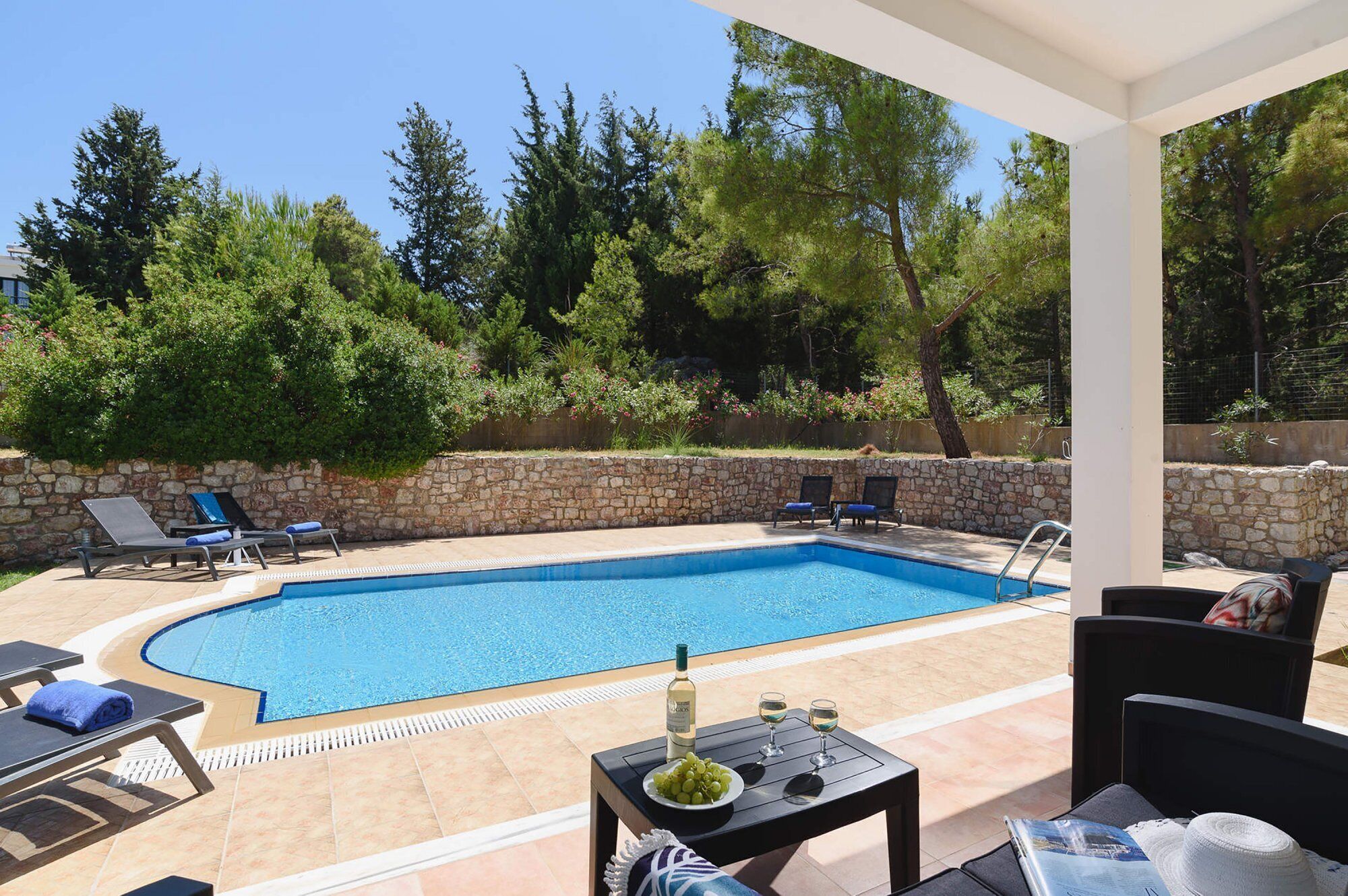 Une piscine avec des chaises longues, une table avec une bouteille et des verres, et un mur en pierre.