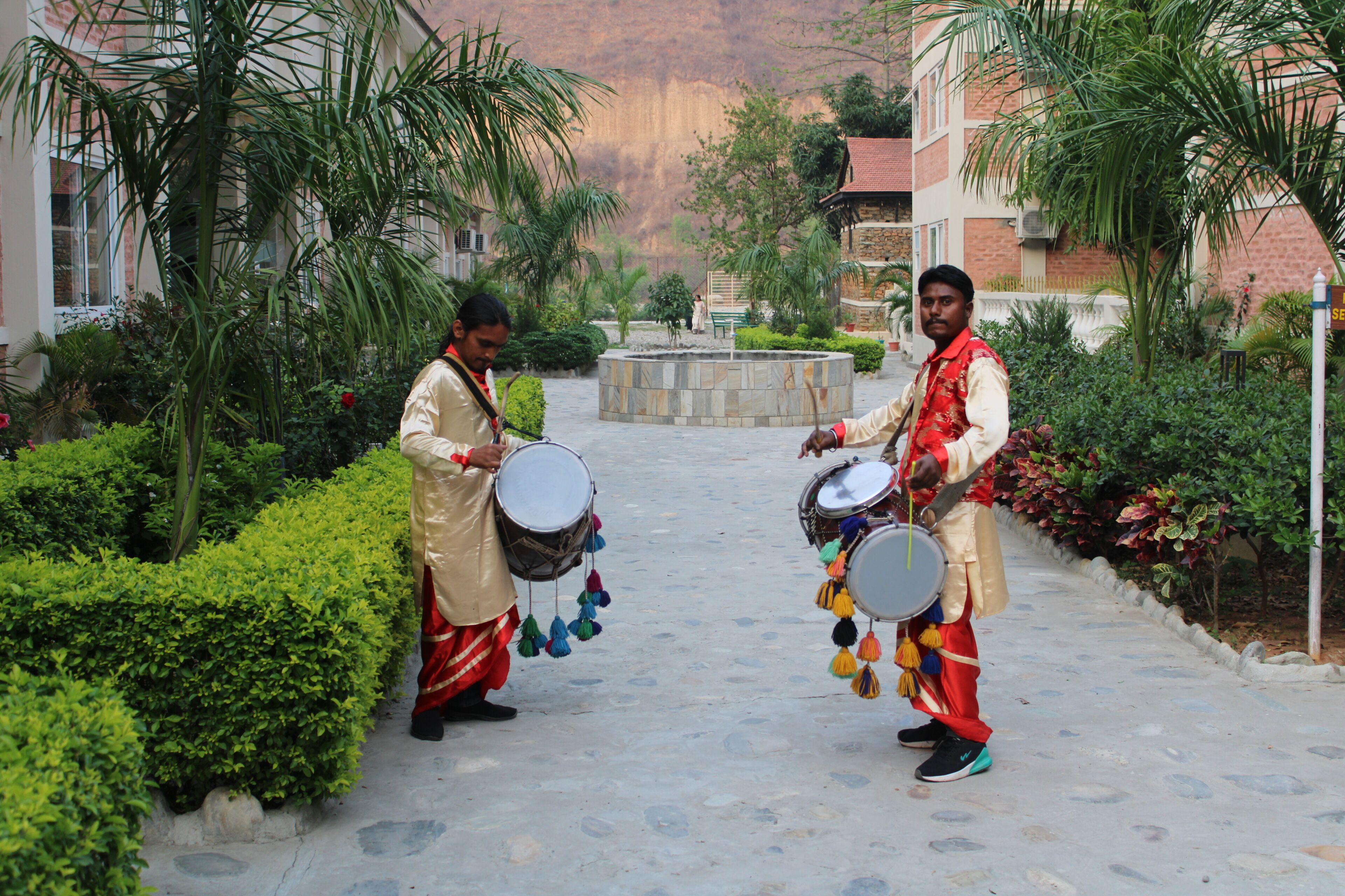 Two drummers in a courtyard with a fountain, surrounded by greenery and buildings.